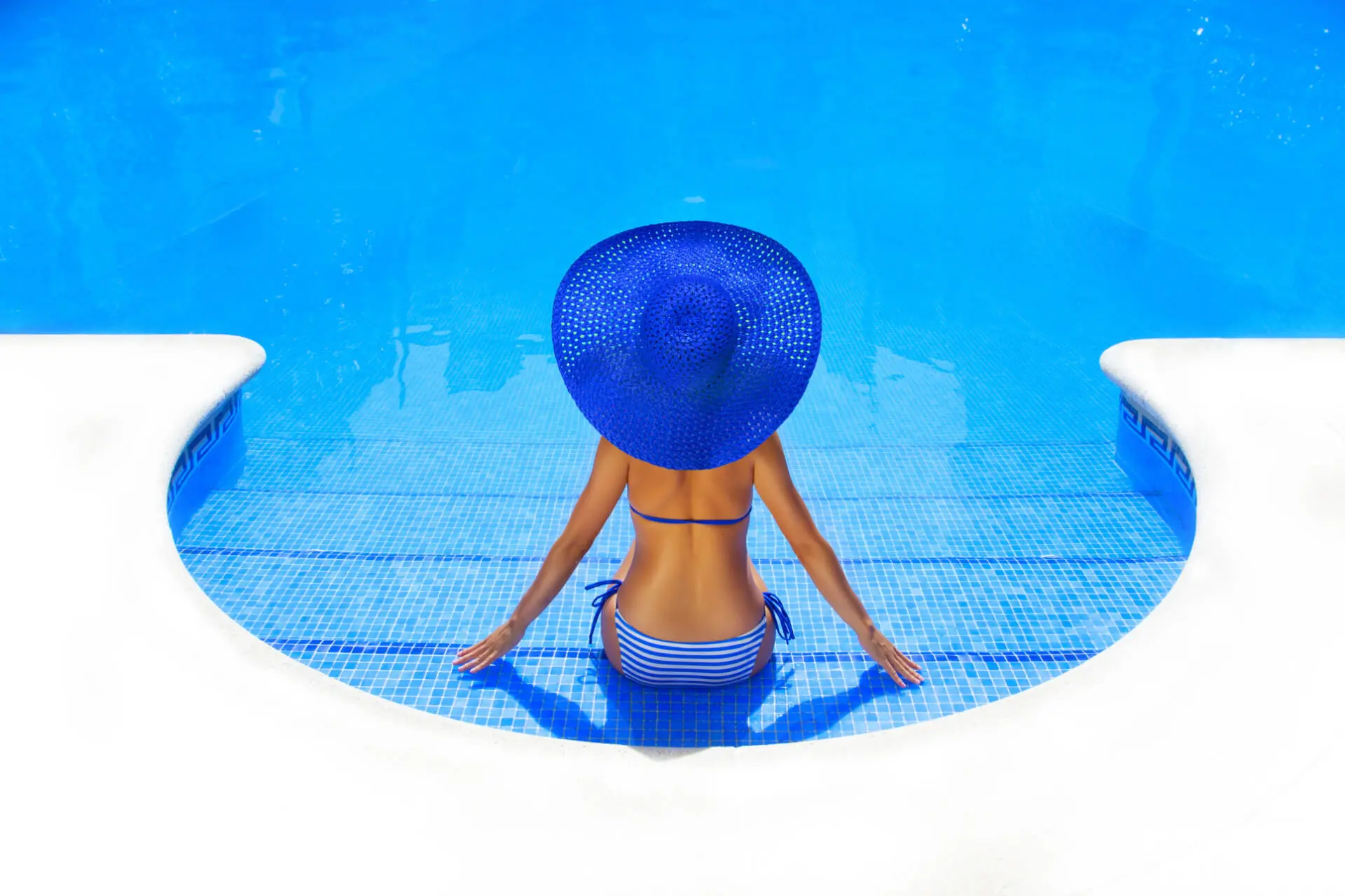 Donna con cappello seduta sulle scale di accesso all'acqua cristallina di una piscina.